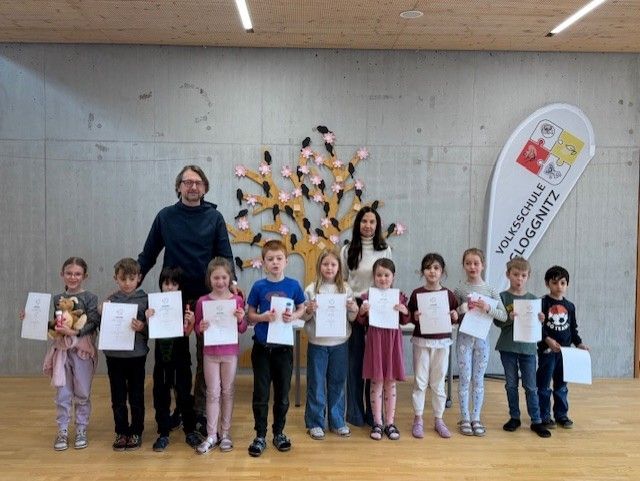 Eine Gruppe von Kindern mit Urkunden steht in einem Raum. Dahinter befinden sich ein Baum und ein Banner an der Wand. Ein Erwachsener steht vorne.