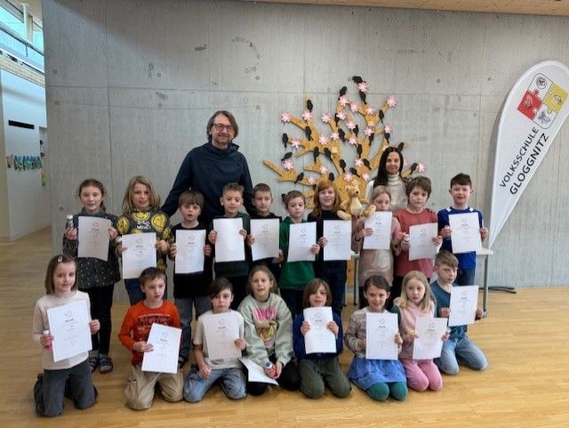 Eine Gruppe von Kindern und zwei Erwachsenen posiert für ein Foto mit Urkunden. Hinter ihnen steht ein Baum mit rosa Blüten.