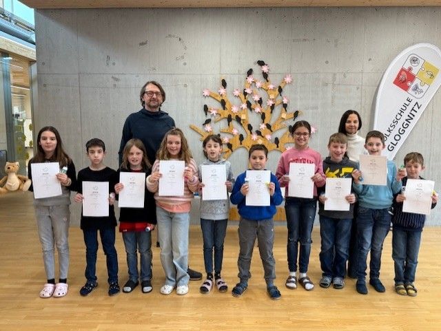 Gruppe von Kindern mit Urkunden posiert für ein Foto vor einer Wand mit einem Baum und Logo.