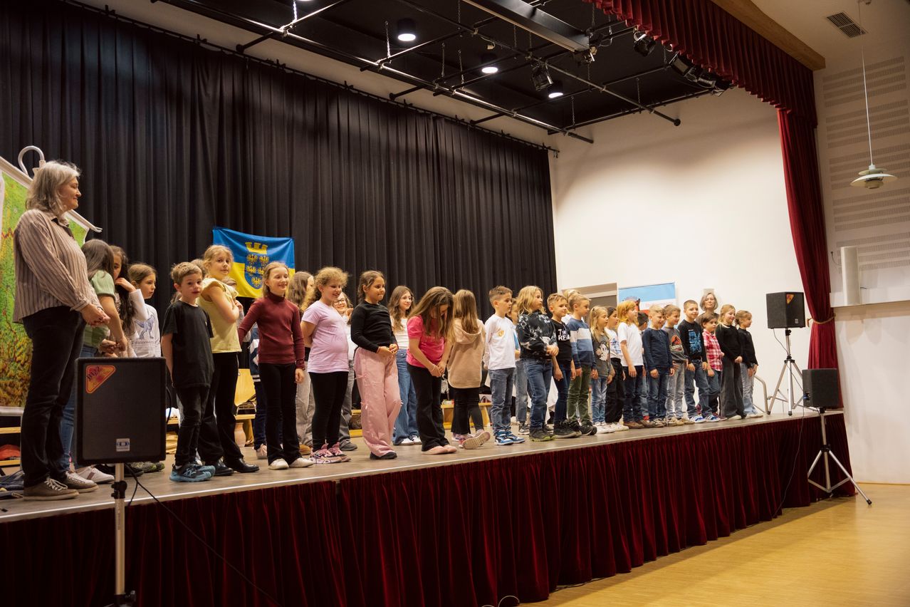 Eine Gruppe von Kindern steht auf einer Bühne, wahrscheinlich bei einer Schulaufführung oder Veranstaltung. Die Bühne ist mit einem roten Vorhang bedeckt.