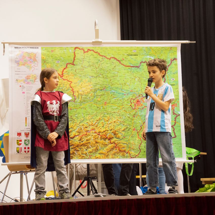 Ein Junge steht mit einem Mikrofon auf der Bühne, gekleidet in einem Trikot. Neben ihm steht ein Mädchen in einem Kostüm neben einer großen Karte. Sie präsentieren wahrscheinlich etwas, mit einer großen Karte im Hintergrund.