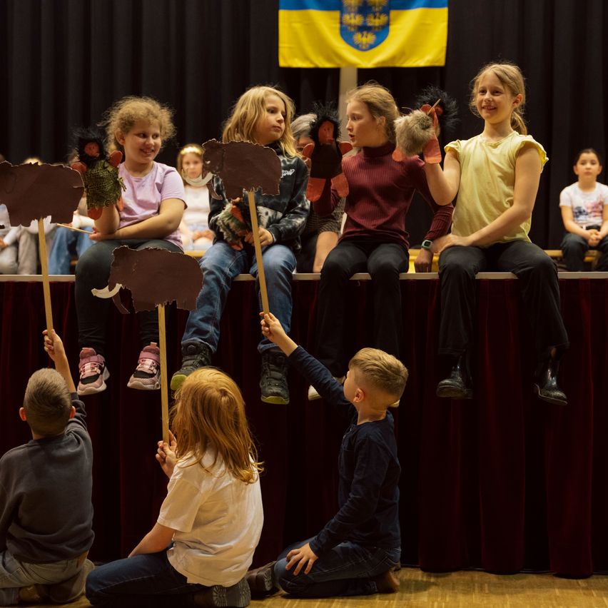 Kinder sitzen auf der Bühne und halten Puppen, wobei ein Kind eine Hand hebt. Dahinter hält ein weiteres Kind ein Schild. Im Hintergrund sind eine Flagge und ein Publikum zu sehen.