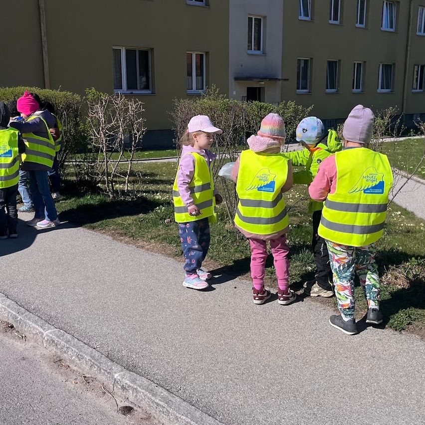 Kinder in Neonwesten stehen auf einem Bürgersteig, schauen auf einen Busch, in der Nähe eines Gebäudes mit vielen Fenstern.