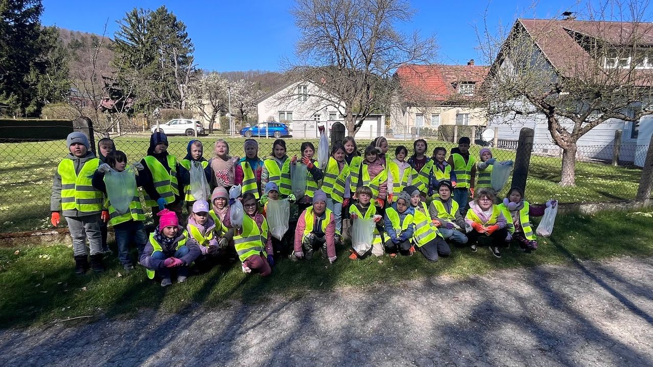 Eine Gruppe von Kindern in Warnwesten posiert für ein Foto in der Nähe eines Zauns, mit Häusern und Bäumen im Hintergrund. Einige halten Beutel.