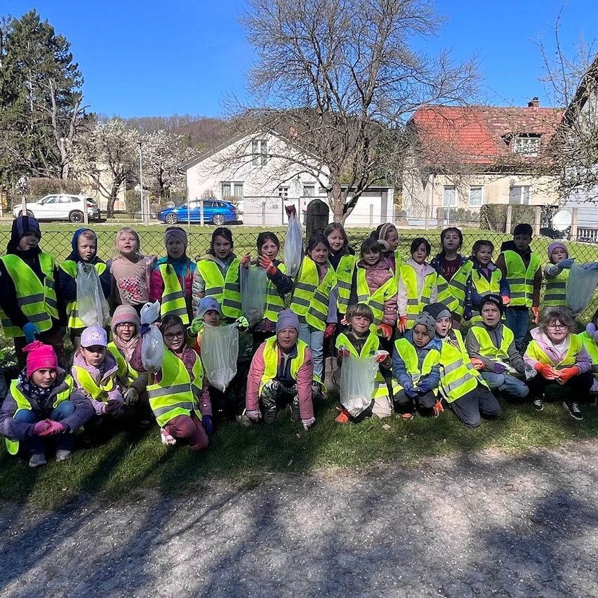 Eine Gruppe von Kindern in Warnwesten posiert für ein Foto in der Nähe eines Zauns, mit Häusern und Bäumen im Hintergrund. Einige halten Beutel.