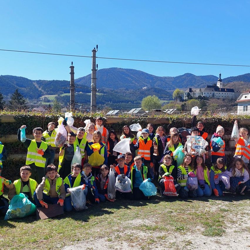 Eine Gruppe von Kindern mit Sicherheitswesten und Mülltüten posiert für ein Foto. Im Hintergrund sind Berge zu sehen.