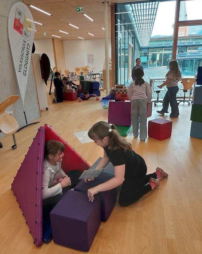 Zwei Mädchen spielen mit Kissen in einem Spielbereich, eine liest ein Buch, während die andere hilft. Andere Kinder spielen ebenfalls. Eine Uhr ist draußen sichtbar.