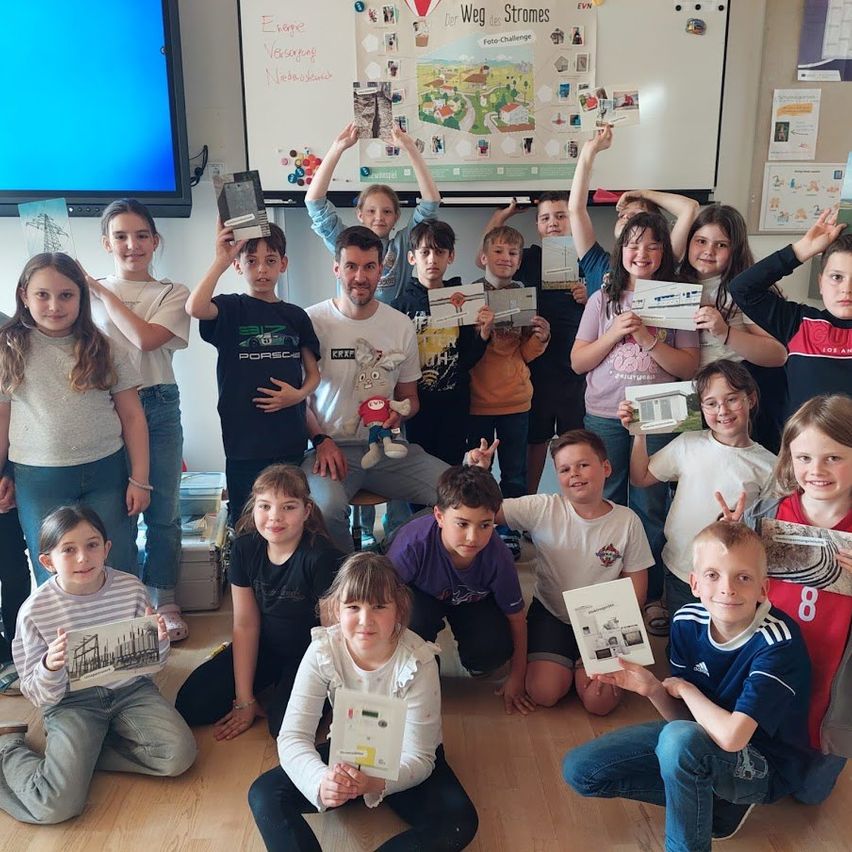 Eine Gruppe von Kindern und einem Mann posiert für ein Foto mit verschiedenen Papieren in einer Klassenzimmerumgebung, mit einer Tafel im Hintergrund.