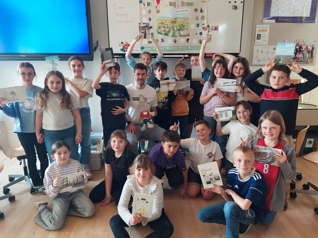 Eine Gruppe von Kindern und einem Mann posiert für ein Foto mit verschiedenen Papieren in einer Klassenzimmerumgebung, mit einer Tafel im Hintergrund.