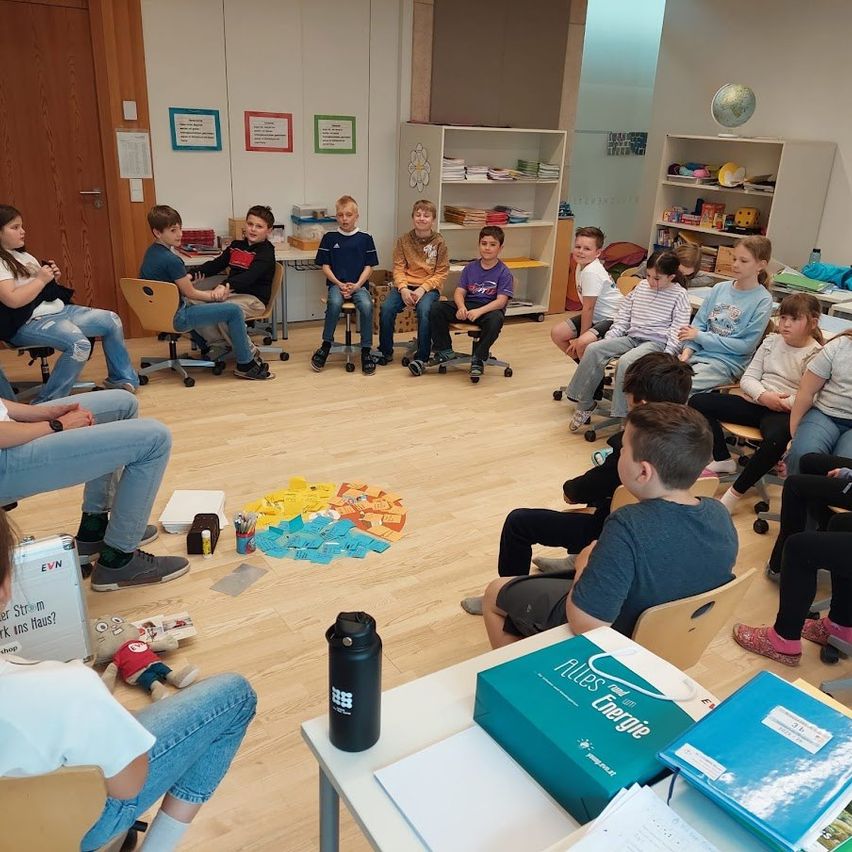 Eine Gruppe von Kindern und Erwachsenen sitzt in einem Klassenzimmer mit Holzboden und weißen Wänden in einem Kreis, ein Globus und Bücher sind sichtbar. Papiere liegen auf dem Boden verstreut.