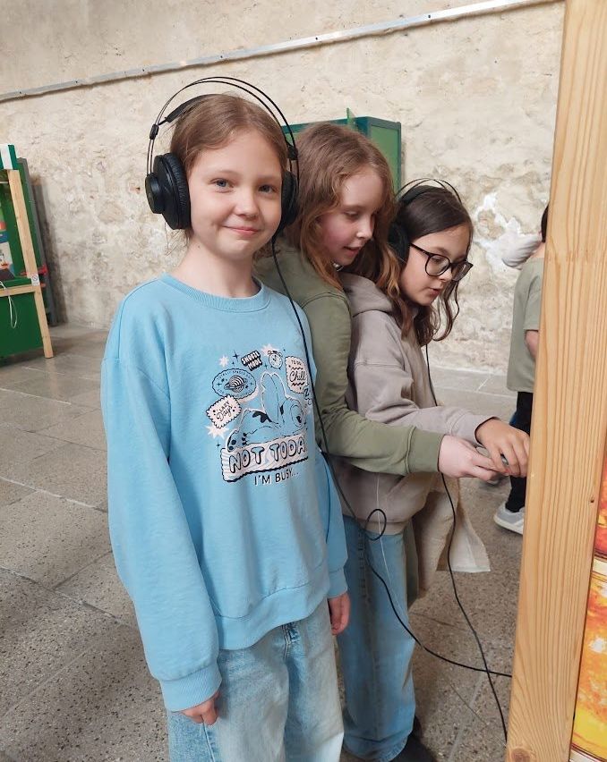 Drei junge Mädchen tragen Kopfhörer und lächeln. Das Mädchen in der Mitte schaut etwas vor sich an, während das Mädchen auf der linken Seite etwas auf ihrer linken Seite anschaut. Das Mädchen auf der rechten Seite schaut nach unten.
