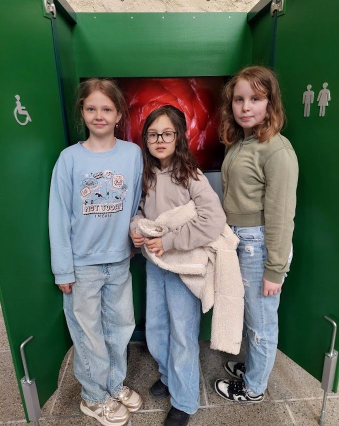 Drei Mädchen stehen zusammen vor einer grünen Kabine, eines hält ein Handtuch. Ein Mädchen trägt eine Brille und ein blaues Sweatshirt mit einer Grafik, die anderen tragen grüne und beige Oberteile. Hinter ihnen befindet sich eine grüne Wand mit einem roten Grafik und einem Rollstuhlsymbol.