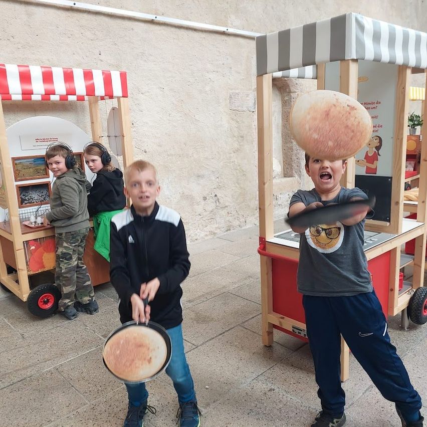 Drei Jungen spielen mit Pfannkuchen an einem Imbissstand, einer balanciert einen Pfannkuchen auf seinem Kopf. In der Nähe hilft ein Mädchen an einem anderen Imbissstand.