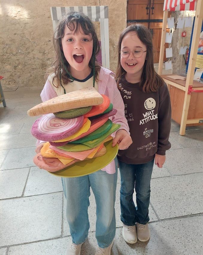 Zwei junge Mädchen halten einen bunten Holzsandwich, eine lacht breit, die andere trägt eine Brille. Dahinter steht eine Holzstruktur mit angehefteten Papieren.