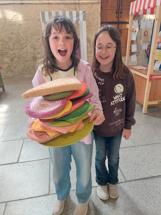 Zwei junge Mädchen halten einen bunten Holzsandwich, eine lacht breit, die andere trägt eine Brille. Dahinter steht eine Holzstruktur mit angehefteten Papieren.