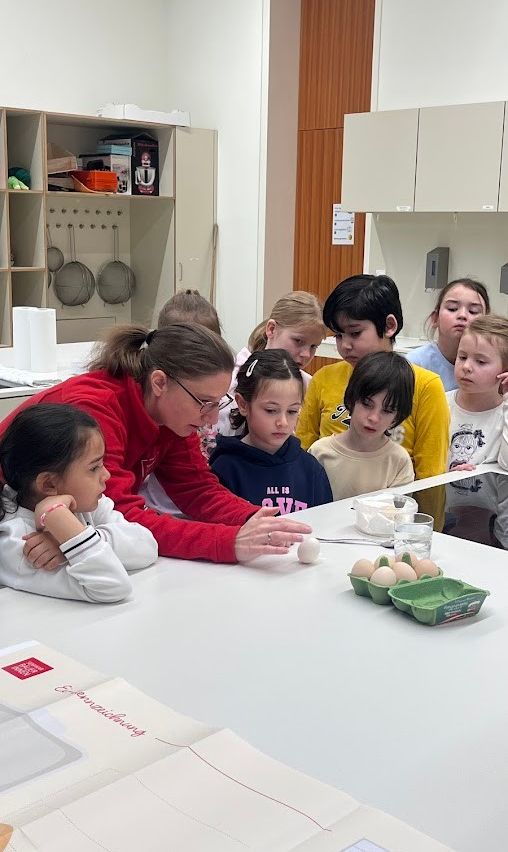 Eine Gruppe von Kindern sitzt mit einer Frau in einer roten Jacke an einem Tisch. Sie schauen ein Ei an und es gibt Eier in einem Karton auf dem Tisch.