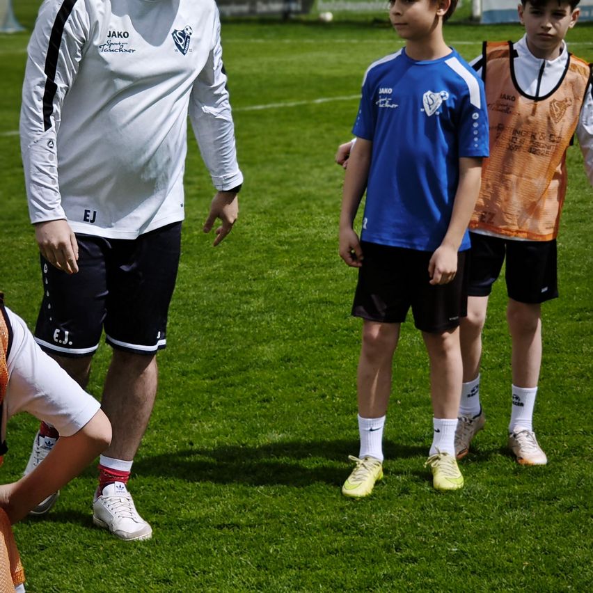 Ein Fußballtrainer in einem weißen Trikot steht auf einem Feld mit zwei Jungen in blauen und orangefarbenen Uniformen. Das Feld ist grün und der Himmel ist klar.