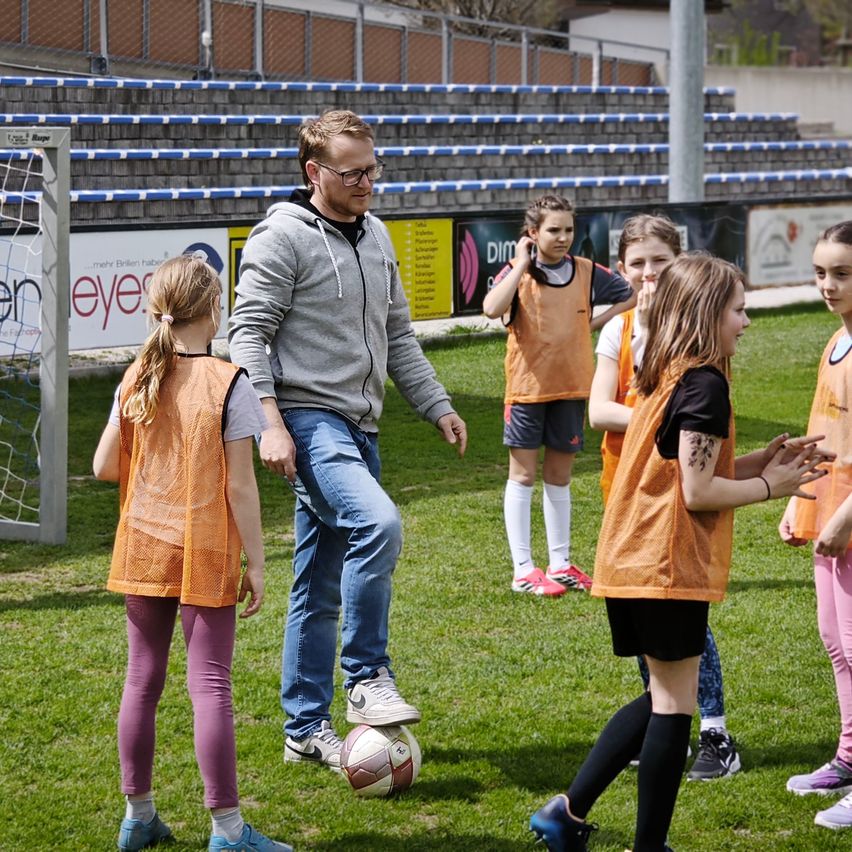 Ein Mann in einer grauen Kapuzenjacke unterrichtet junge Mädchen im Fußball auf einem Spielfeld. Hinter ihnen sind leere Tribünen.
