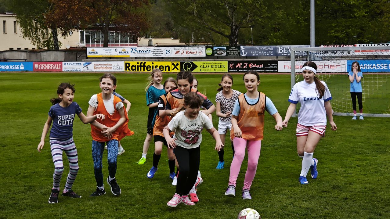 Mehrere Mädchen sind auf einem Fußballfeld und spielen Fußball. Einige tragen Turnschuhe, andere Stiefel. Sie tragen bunte Trikots und lachen und lächeln. Ein Fußball liegt auf dem Boden.