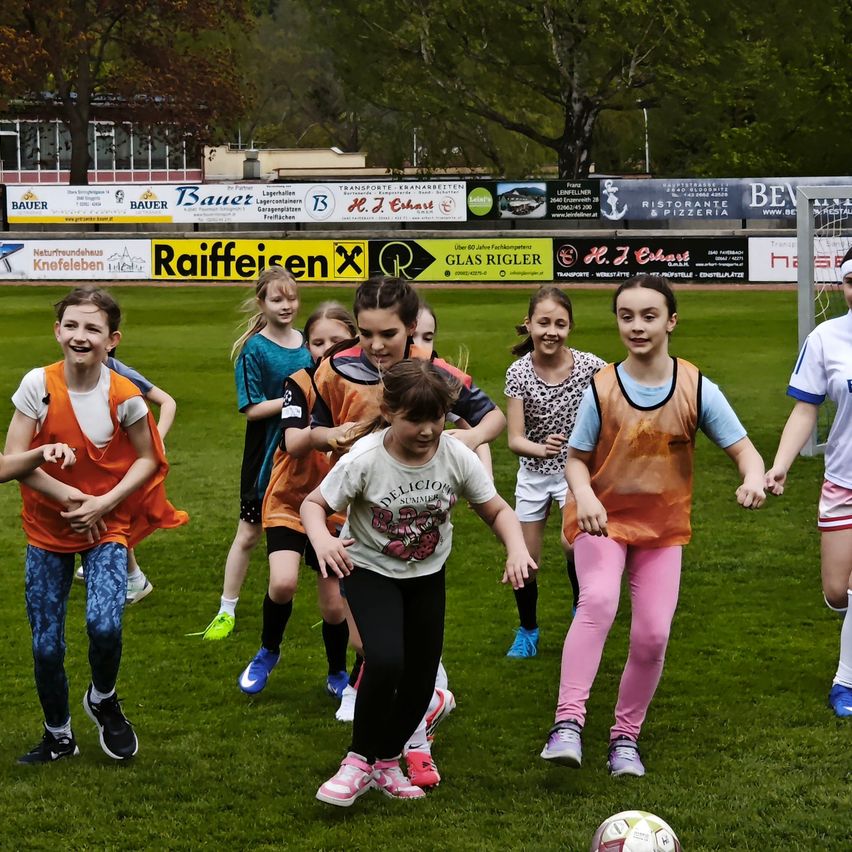 Mehrere Mädchen sind auf einem Fußballfeld und spielen Fußball. Einige tragen Turnschuhe, andere Stiefel. Sie tragen bunte Trikots und lachen und lächeln. Ein Fußball liegt auf dem Boden.