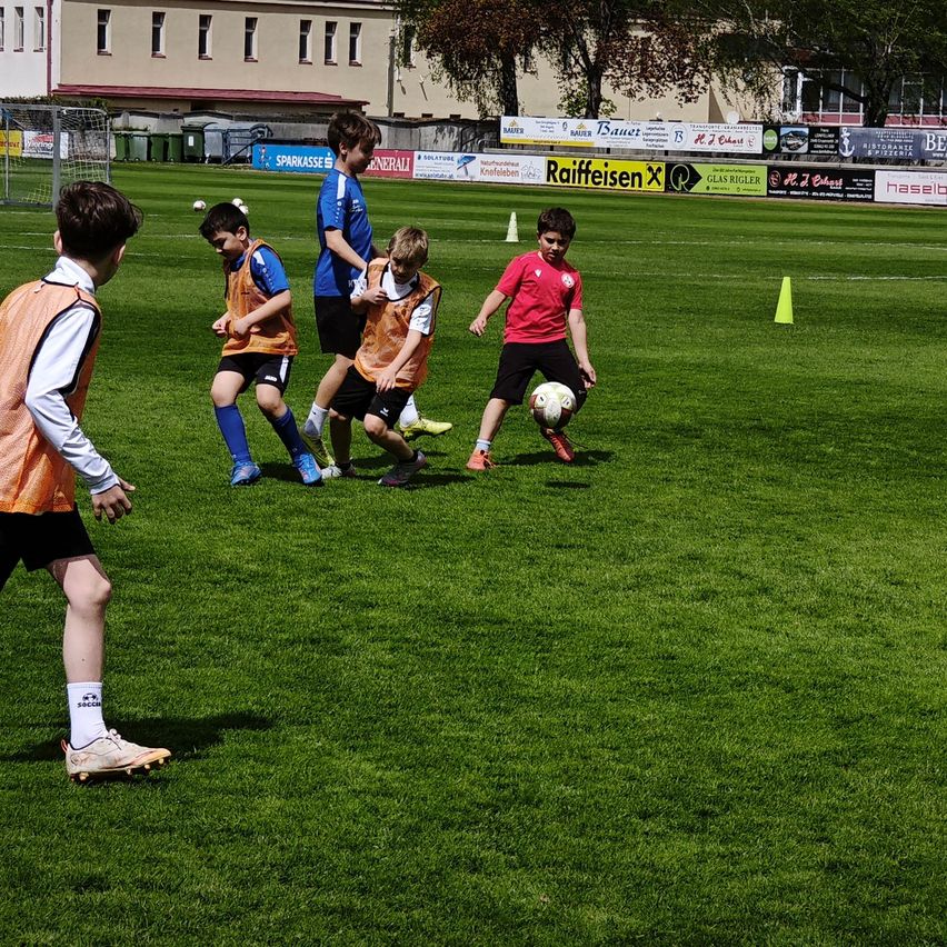 Eine Gruppe von Kindern spielt Fußball auf einem grünen Feld. Sie tragen orangefarbene Westen. Ein Kind in einem roten Shirt kickt den Ball.
