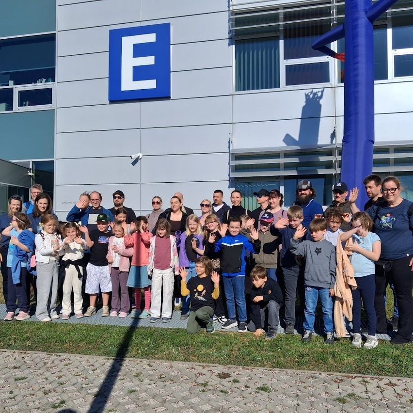 Eine Gruppe von Menschen steht vor einem Gebäude mit einem blauen E-Logo. Einige winken und posieren für ein Foto.