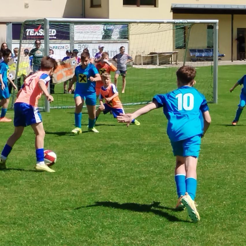 Eine Gruppe von Kindern spielt Fußball auf einem Feld. Spieler Nummer 10 versucht, einen Schuss zu blocken. Der Ball liegt neben einem Jungen auf dem Boden.