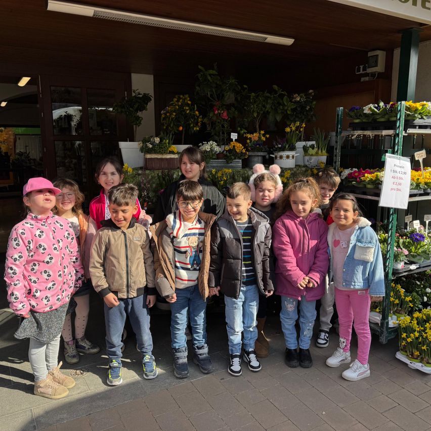 Eine Gruppe von Kindern steht vor einem Blumenladen und lächelt für die Kamera. Hinter ihnen sind Topfpflanzen und Blumen ausgestellt.