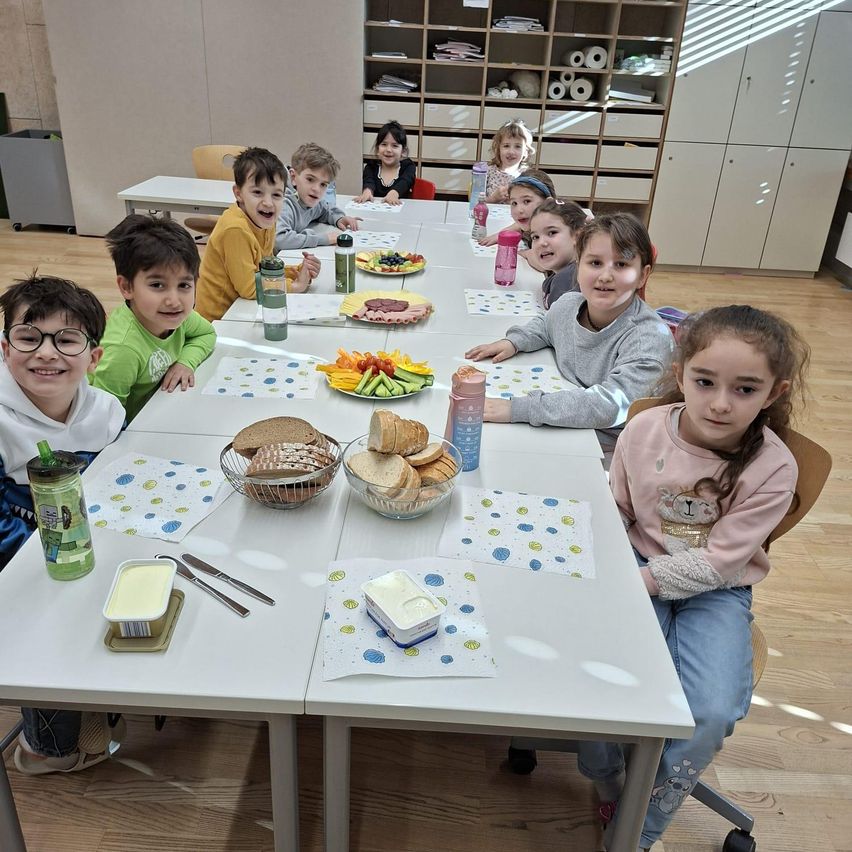 Eine Gruppe von Kindern sitzt an Tischen mit Tellern voller Speisen, Wasserflaschen und Papierservietten. Ein Kind trägt eine Brille. Sie sind in einem Klassenzimmer mit Holzböden.