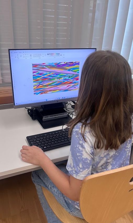 Ein Mädchen sitzt an einem Schreibtisch mit einem Computer und konzentriert sich auf ein farbenfrohes, abstraktes Design auf dem Bildschirm. Sie trägt ein blaues T-Shirt.