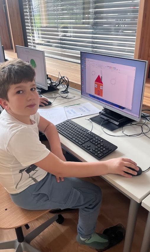 Ein junger Junge sitzt an einem Schreibtisch, konzentriert auf einen Computerbildschirm, der ein Haus-Zeichnen zeigt. Papiere und eine Tastatur sind auf dem Schreibtisch.