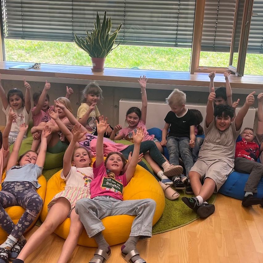 Eine Gruppe junger Kinder, die auf Bohnensäcken drinnen sitzen, ihre Hände heben, lächeln und in die Kamera schauen.