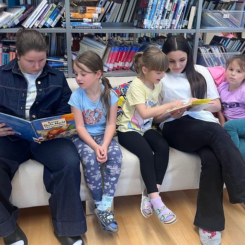 Zwei Frauen und vier junge Mädchen sitzen gemeinsam auf einer Couch in einer Bibliothek. Die Frauen lesen den Mädchen vor. Die Mädchen sehen interessiert in die Bücher.