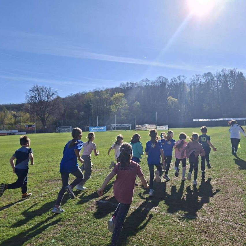 Eine Gruppe von Kindern läuft in einer Reihe auf einem grasigen Feld unter einem hellen Himmel mit entfernten Bäumen. Einige tragen Turnschuhe, andere Stiefel.