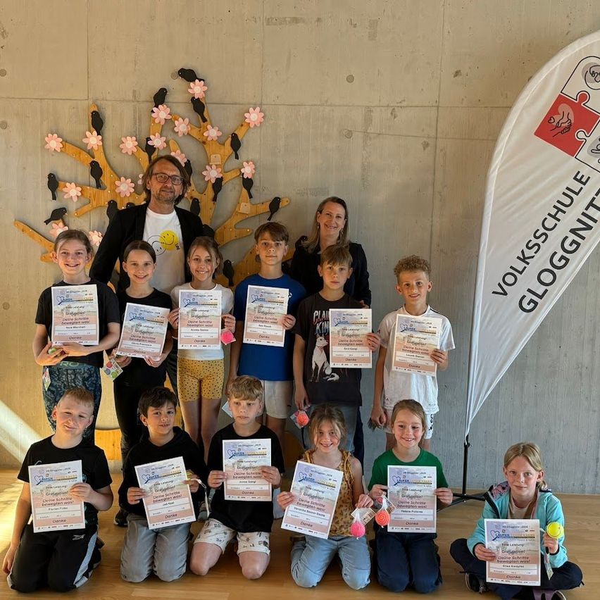 Eine Gruppe von Kindern, Erwachsenen und einer Fahne stehen in einem Raum, halten Urkunden. Die Fahne steht 'Volksschule GLOGGNITZ'. Die Kinder lächeln und posieren für ein Foto.