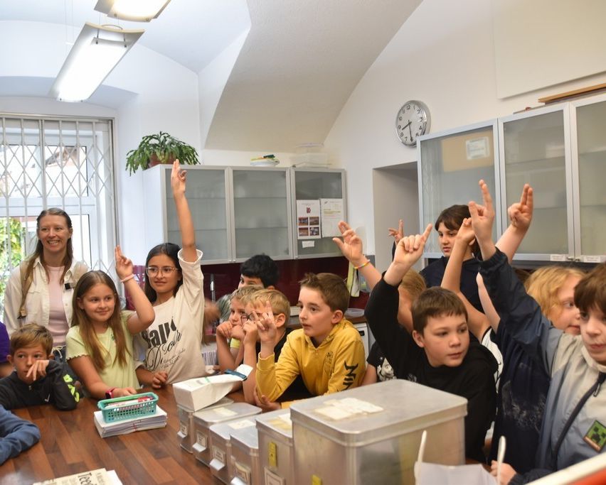 Eine Gruppe von Kindern und eine Frau sitzen an einem Tisch. Die Kinder heben ihre Hände. Hinter ihnen ist eine Wand mit Schränken, einem Fenster mit Gitter und einer Uhr.