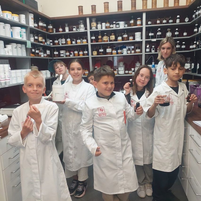 Eine Gruppe von Kindern in Laborkitteln steht in einem Labor und hält Bechergläser. Sie posieren für ein Foto. Regale mit zahlreichen Flaschen befinden sich hinter ihnen.
