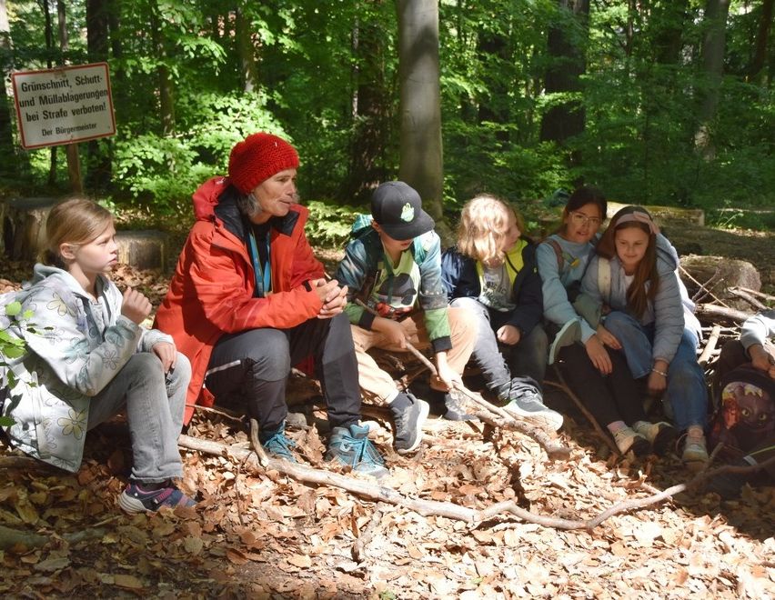 Eine Gruppe von Kindern sitzt auf einem Haufen Laub in einem Wald, eine Frau in einer roten Mütze lehrt sie.