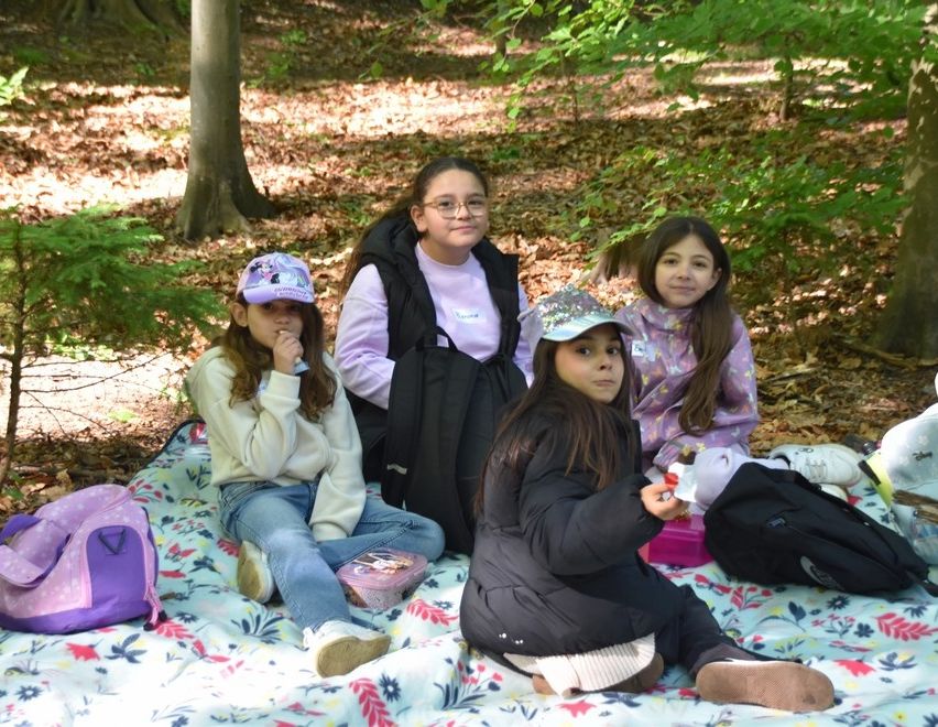 Fünf Mädchen sitzen auf einer Decke im Wald, umgeben von gefallenen Blättern. Eines trägt eine Brille und einen Rucksack. Eine andere hat eine rosa Lunchbox. Taschen und Schuhe sind herum verstreut.