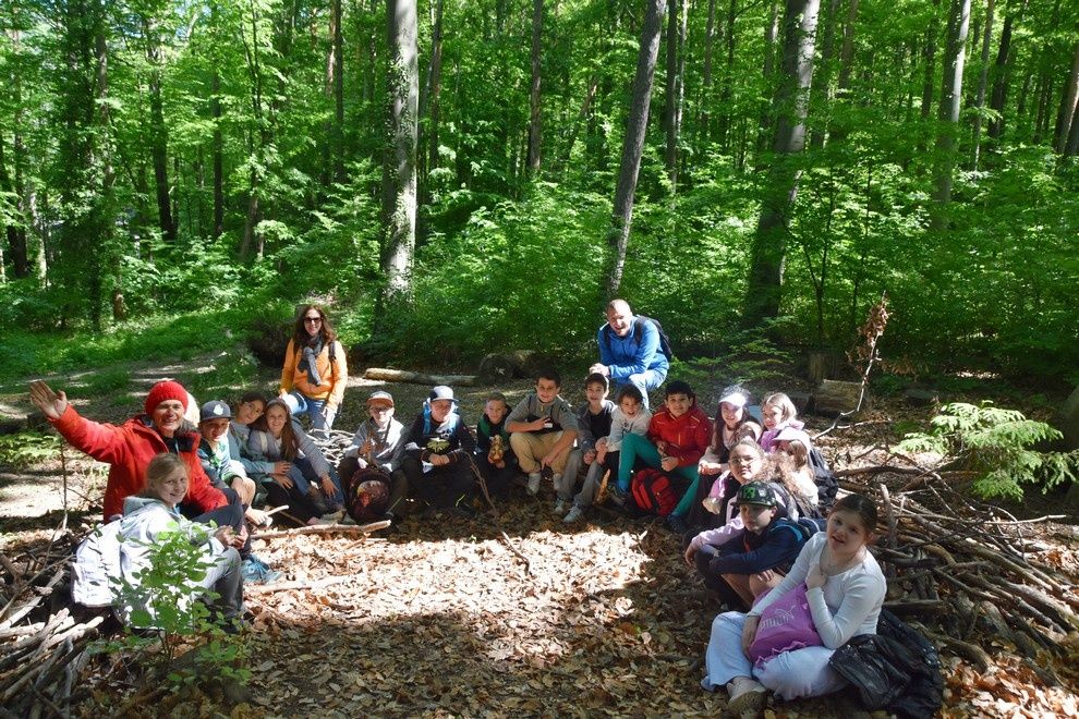 Eine Gruppe von Menschen, wahrscheinlich Schüler, versammelt sich in einer Waldlichtung. Sie sitzen und stehen in einem Halbkreis, einige mit Rucksäcken und Mützen.
