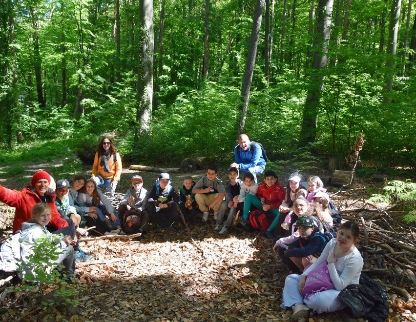Eine Gruppe von Menschen, wahrscheinlich Schüler, versammelt sich in einer Waldlichtung. Sie sitzen und stehen in einem Halbkreis, einige mit Rucksäcken und Mützen.