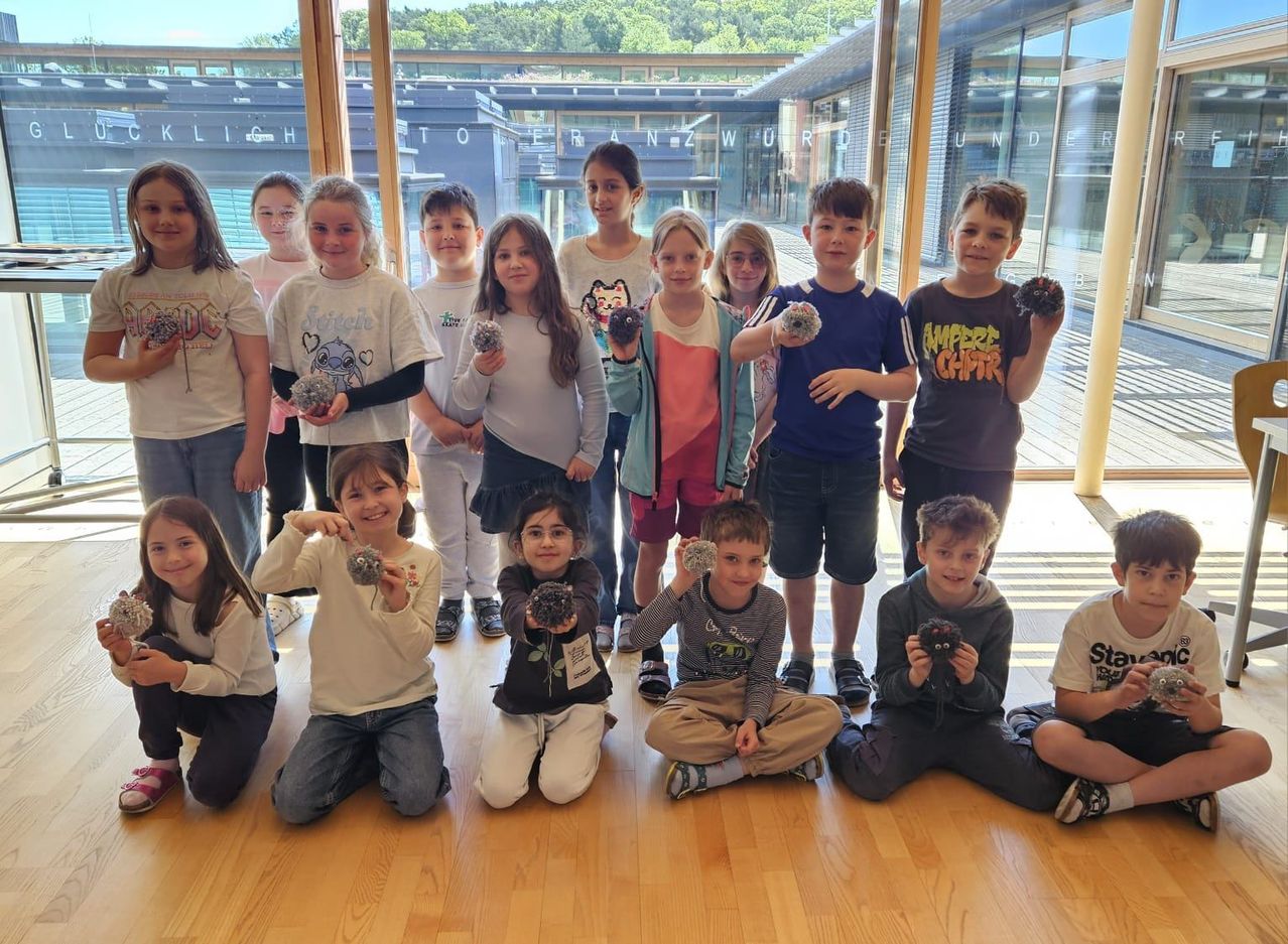 Eine Gruppe von Kindern in Freizeitkleidung posiert drinnen und hält dekorative Objekte. Hinter ihnen ist ein modernes Gebäude mit Glasfenstern zu sehen.
