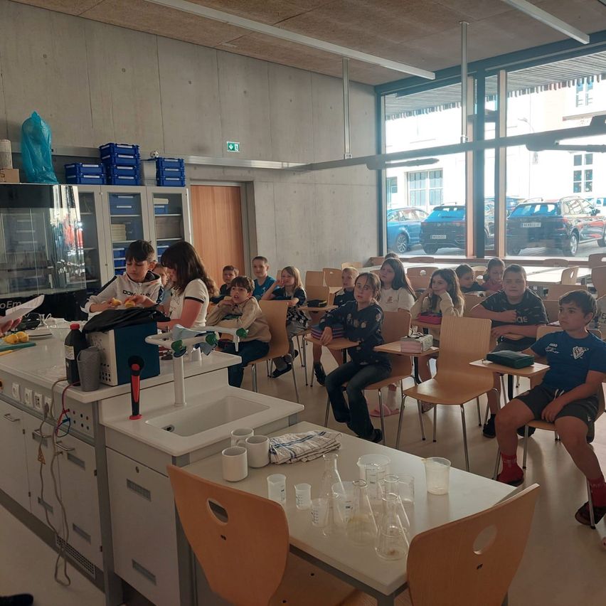 Ein Klassenzimmer mit Schülern an Tischen; eine Lehrerin bereitet ein Experiment vor. Der Raum ist gut beleuchtet mit großen Fenstern. Ein Waschbecken und ein Tisch halten verschiedene wissenschaftliche Geräte.
