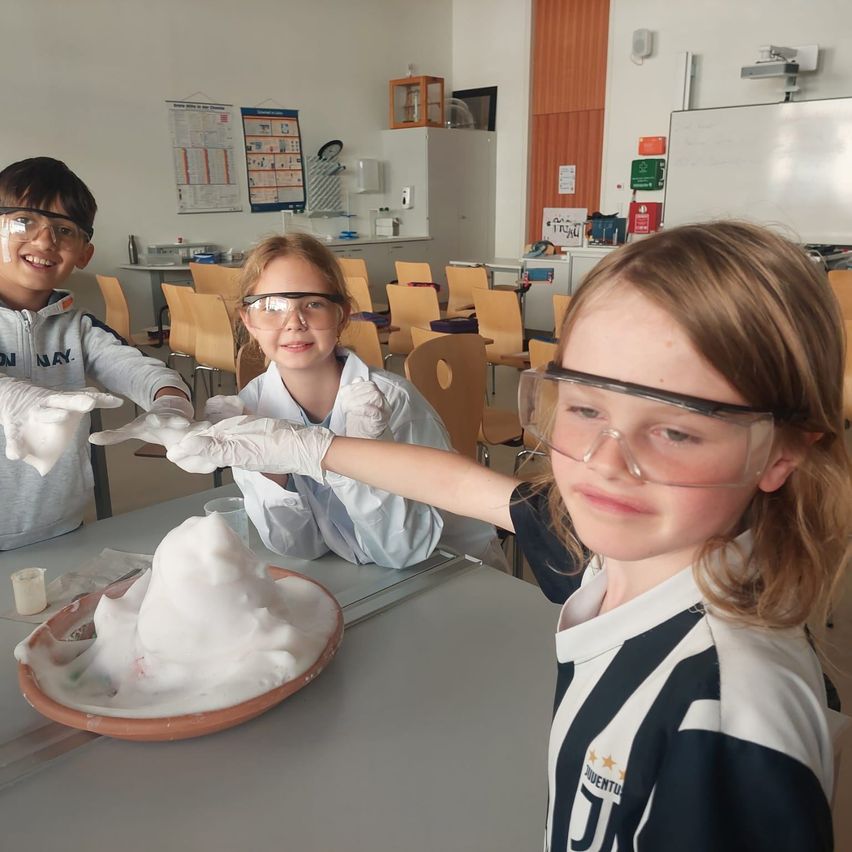 Drei Kinder sind in einem Klassenzimmer, tragen Schutzbrillen und Handschuhe. Sie spielen mit einem Vulkanexperiment auf einem Tisch.