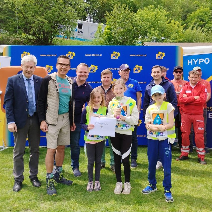 Eine Gruppe von Erwachsenen und Kindern posiert für ein Foto vor einem blauen Banner. Die Kinder halten Urkunden und tragen Sicherheitswesten. Zu den Erwachsenen gehören ein Mann in Anzug und Krawatte, ein Mann mit Brille und weitere Personen.
