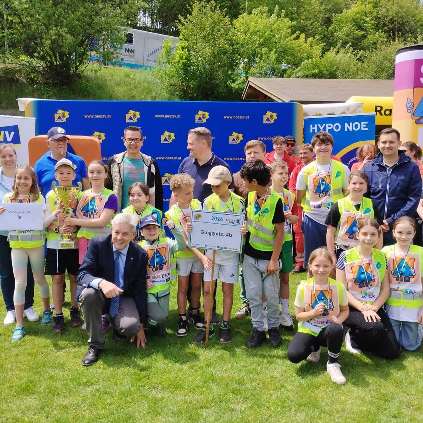 Eine Gruppe von Kindern und Erwachsenen posiert für ein Foto auf einem Rasenfeld mit Bannern dahinter. Die Kinder tragen Sicherheitswesten und halten ein Zertifikat. Ein Mann kauert sich im Vordergrund.