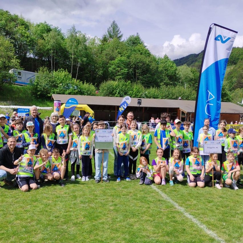 Eine Gruppe von Kindern und Erwachsenen in grünen Westen posiert für ein Foto auf einem Rasenfeld, mit einer Fahne und einem Gebäude im Hintergrund.