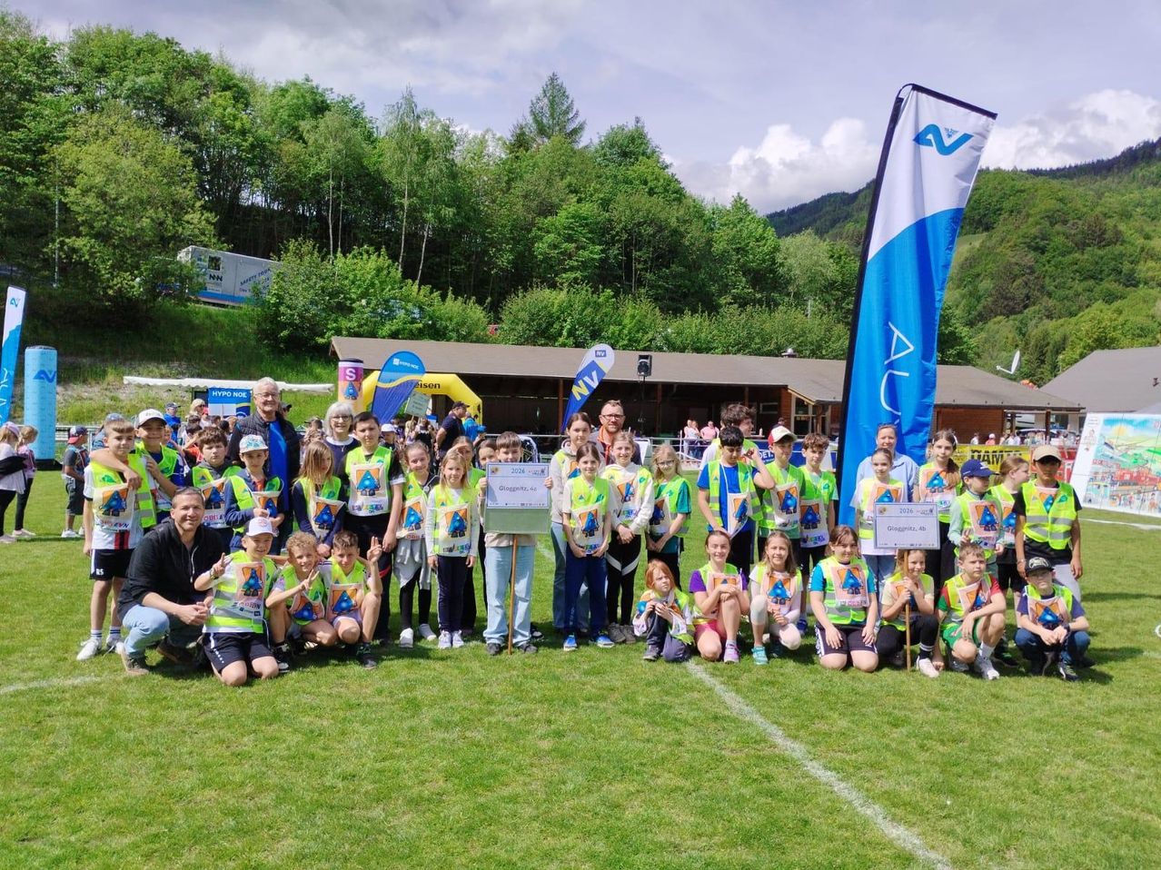 Eine Gruppe von Kindern und Erwachsenen in grünen Westen posiert für ein Foto auf einem Rasenfeld, mit einer Fahne und einem Gebäude im Hintergrund.