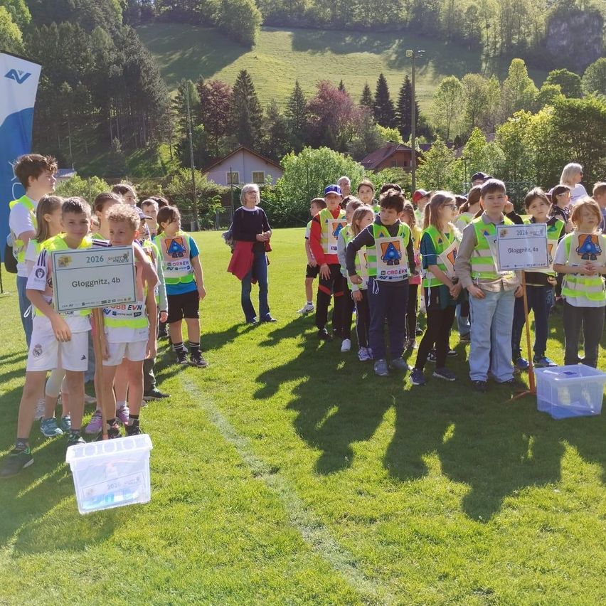 Eine Gruppe von Kindern in gelben Westen hält Pokale auf einem Rasenfeld hoch. Sie stehen in einer Reihe. Dahinter stehen einige Menschen und schauen zu. Im Hintergrund sind Bäume und Häuser zu sehen.