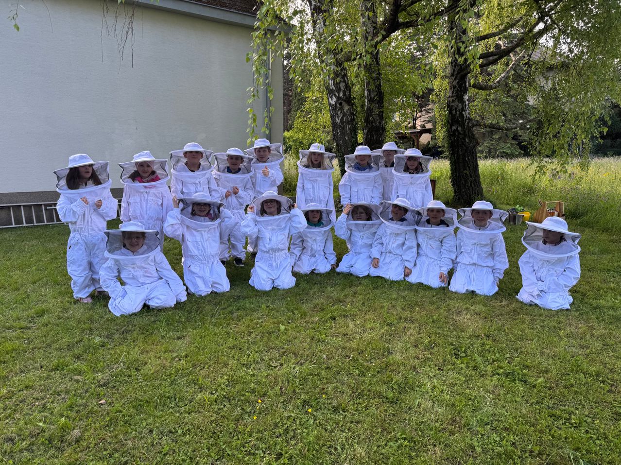 Eine Gruppe von Imkern in voller Schutzausrüstung posiert für ein Foto auf einer Rasenfläche in der Nähe von Bäumen und einem Gebäude.